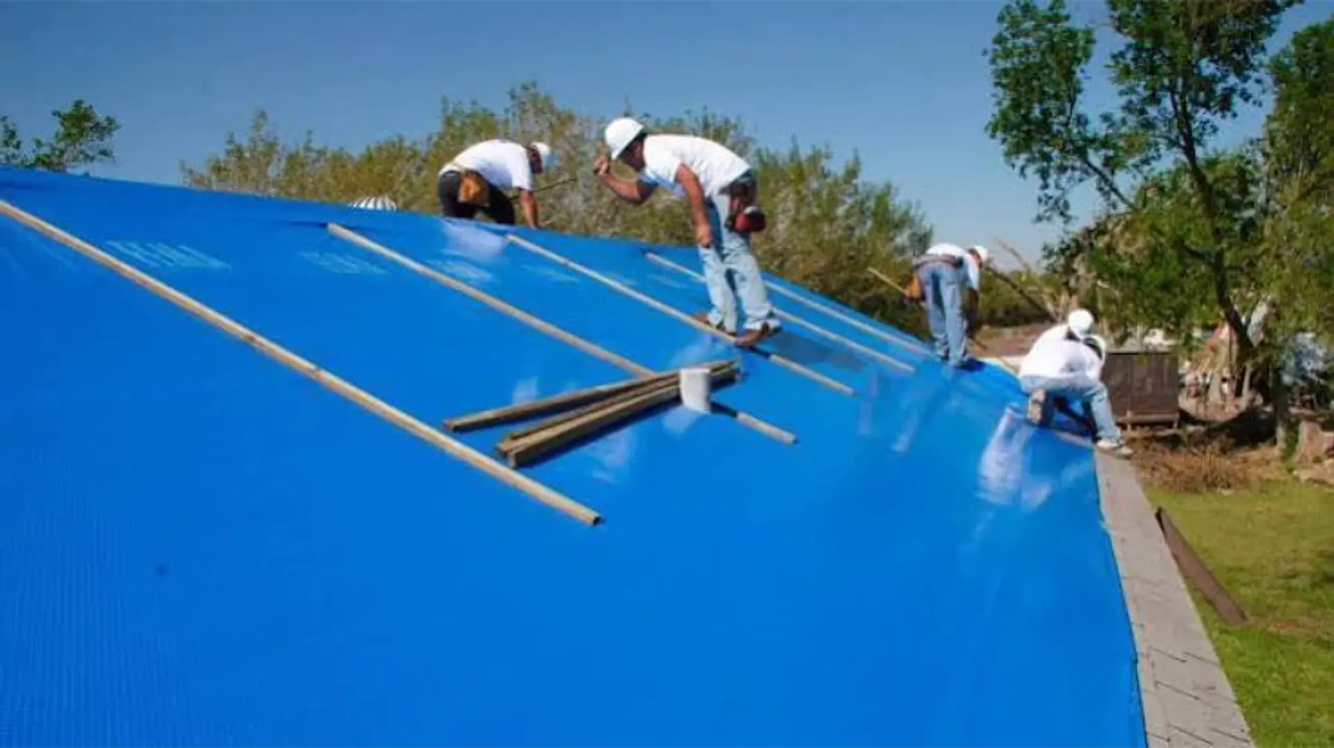 Crew securing a blue emergency tarp over a damaged roof with wooden battens
